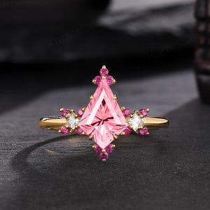 May include: A gold ring featuring a large, kite-shaped pink gemstone as the centerpiece. The ring is adorned with smaller pink and white gemstones, creating a symmetrical design. The ring is set against a dark background, highlighting its sparkle.