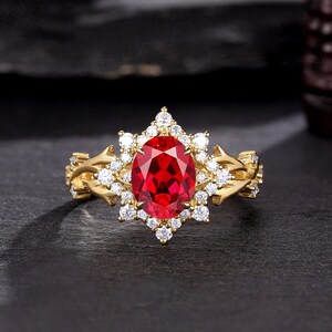 Può includere: Un anello d'oro con una grande pietra ovale rossa circondata da pietre bianche più piccole. L'anello ha un design delicato e intricato con un motivo floreale.