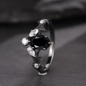 May include: Black engagement ring with a large oval black gemstone surrounded by smaller white gemstones. The ring band is black and has a delicate floral design.
