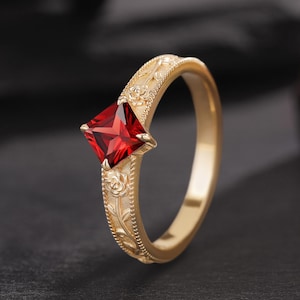 May include: A gold ring featuring a square-cut deep red gemstone set in the center. The band is crafted with a detailed relief pattern of roses and vines along the outer surface, framed by thin beaded edges that run along the entire circumference.