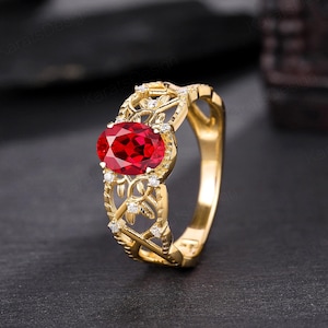 Puede incluir: Un anillo de oro con una piedra ovalada roja rodeada de piedras blancas más pequeñas. El anillo tiene un delicado diseño floral.