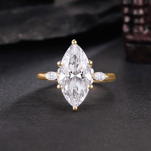 Op de afbeelding: Een gouden verlovingsring met een opvallende marquise-geslepen diamant. De ring is versierd met kleinere, peervormige diamanten. De band is goudkleurig, tegen een donkere achtergrond. Het ontwerp van de ring is elegant en klassiek.