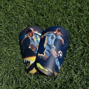 Könnte beinhalten: Ein Paar schwarze Schienbeinschoner mit einem blauen und weißen Fußballspieler-Design. Die Schienbeinschoner zeigen einen Fußballer, der einen Ball tritt, und die Nummer 10 auf dem Trikot.