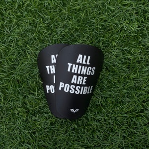 Könnte beinhalten: Zwei schwarze Schienbeinschoner mit weißem Text, der "All things are possible" lautet, und einem Logo eines stilisierten "F" mit Flügeln.