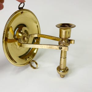 May include: A vintage brass wall-mounted candle holder. The candle holder features a circular backplate, an adjustable arm, and a cup-shaped holder for the candle. The brass has a polished finish, reflecting light. The design is classic and elegant.