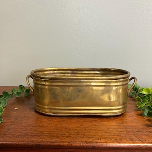 May include: An oval-shaped brass planter with a polished metallic finish, featuring two small handles on opposite sides and a horizontal ribbed design along the exterior walls. The container has a wide opening and a flat base.
