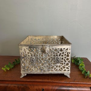 May include: A silver-toned metal decorative box with intricate openwork patterns. The box features a lion's head handle and decorative floral accents. The rectangular box sits on four small feet, adding to its vintage appeal.