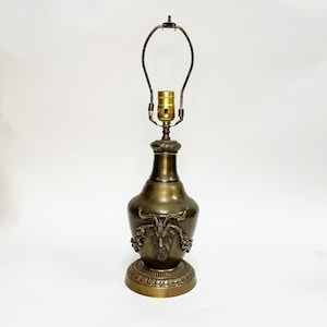 May include: A vintage brass table lamp with a decorative base and a detailed design featuring a ram's head. The lamp has a curved metal shade holder and a bulb socket. The lamp is approximately 20 inches tall.