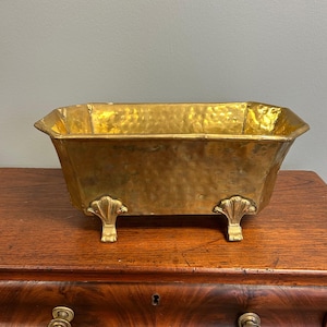 May include: A rectangular brass planter with a hammered texture and a polished gold-tone finish. The vessel features four decorative feet shaped like scallop shells, providing a raised base for the container's wide, open-top basin design.