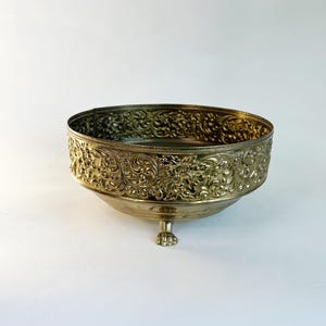 May include: A gold-colored, ornate bowl with a detailed floral pattern around the rim and sides. The bowl has a rounded base and a single, decorative foot. The bowl is likely vintage or antique.
