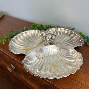 May include: A silver-toned metal serving dish shaped like three conjoined scallop shells. The dish features a central handle sculpted in the form of a small squirrel sitting upright. The surface has a ribbed texture and a metallic finish.