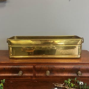 May include: A rectangular brass planter with a hammered finish and rounded corners. The container features a polished metallic surface that reflects light, with small decorative handles attached to each of the two shorter side panels.
