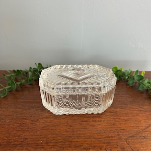May include: A clear, pressed glass trinket box with an octagonal shape. The lid features a geometric diamond pattern in the center, while the sides of the box are decorated with vertical, wavy fluted ridges that catch the light.