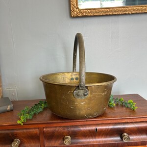 May include: A vintage brass bucket with a curved handle. The bucket has a round body and a slightly flared rim. The brass has a warm, aged patina. The handle is attached with rivets. The bucket is sitting on a wooden surface.