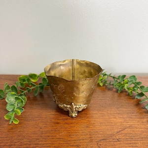 May include: A small, hammered brass bowl with a wavy, scalloped rim and a weathered, aged patina. The bowl features three ornate, decorative feet with intricate leaf-like detailing at the base, providing a stable foundation for the vessel.