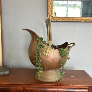 May include: A copper and brass coal scuttle with a handle and decorative accents. The scuttle has a spout and a round base. Greenery is draped over the side. The scuttle is on a wooden surface.