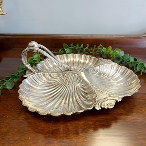 May include: A silver-toned metal serving dish featuring three interconnected shell-shaped compartments. The dish includes a central arched handle with a spherical finial, and the edges of each shell section are decorated with ornate, scalloped detailing.
