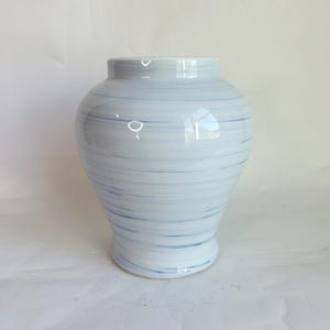May include: A white ceramic vase with a blue swirl pattern. The vase has a wide base that tapers to a narrower neck and a flared rim. The blue lines are evenly spaced and create a textured effect. The vase is a decorative home accent.