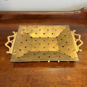 May include: A rectangular brass tray featuring a woven metal lattice pattern across the center. The tray includes two ornate, curved handles on the sides, each attached with visible rivets to the frame of the tray, creating a metallic finish.