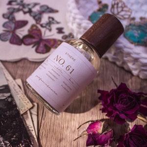 May include: A clear glass bottle of perfume with a brown wooden cap. The bottle is labeled "Ethere No. 61" and lists the notes "middle: mandarin, parchment, rosemary" and "base: rose mousse, plumena".