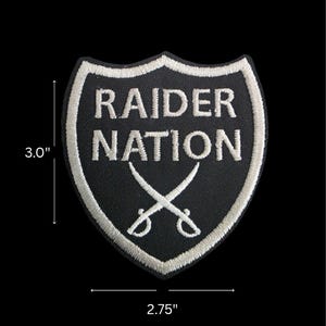 May include: Black and white embroidered patch in a shield shape, measuring 3.0 inches by 2.75 inches. The patch features the words "RAIDER NATION" in white, with two crossed swords below. The border is white.
