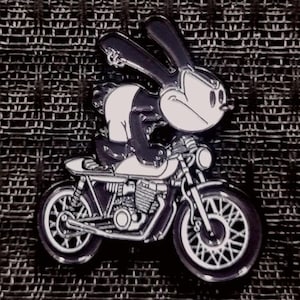 May include: A black and white cartoon rabbit riding a motorcycle. The rabbit is wearing a black leather jacket and has a white face with black eyes and a black nose. The motorcycle is black and has a white seat and a white headlight.