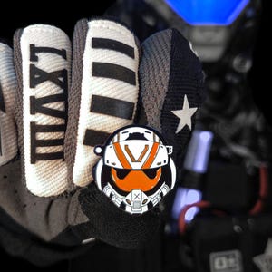 May include: A black, white, and orange pin featuring a stylized helmet design. The pin is held in a black glove, with the Roman numeral "LXVIII" visible on the glove. The background features a futuristic helmet with blue lights.