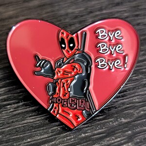 May include: A heart-shaped enamel pin featuring Deadpool in a red and black costume, giving a hand gesture. The pin has the words "Bye Bye Bye!" in white text. The pin is red with a black outline.