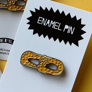 May include: Enamel pin featuring a pretzel with a yellow and brown glaze. The pin is on a white card with the text "Enamel Pin" and "Made by Nilina" in black.