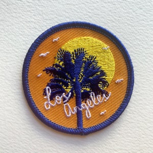 May include: A round, orange embroidered patch featuring a blue palm tree and the text "Los Angeles" in white. The palm tree is silhouetted against a yellow sun.