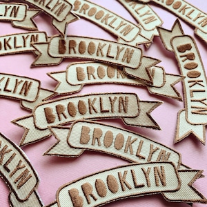May include: A collection of brown embroidered patches with the word "Brooklyn" on them. The patches are shaped like banners and are arranged on a pink background.
