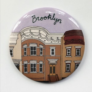 Könnte beinhalten: Ein runder Knopf mit einem violetten Hintergrund und dem Wort "Brooklyn" in grüner Kursivschrift. Der Knopf zeigt eine Cartoon-Illustration von drei braunen Backsteingebäuden mit weißen Verzierungen und Fenstern.