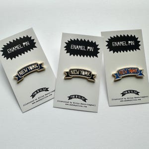 May include: Three enamel pins on white backing cards, each featuring a gold-trimmed banner shape with the text NEW YORK. The pins vary in color, with one dark blue, one black, and one blue with orange lettering, all labeled ENAMEL PIN at the top.