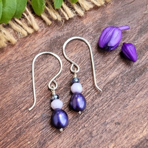 May include: A pair of silver dangle earrings with a purple pearl, a light purple bead, and a dark purple bead on each earring.