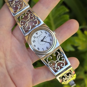 May include: Silver-toned Brighton watch with a white face and black hands. The watch features a rectangular face with a round dial and a silver band with openwork scroll designs. The watch is held in a hand.