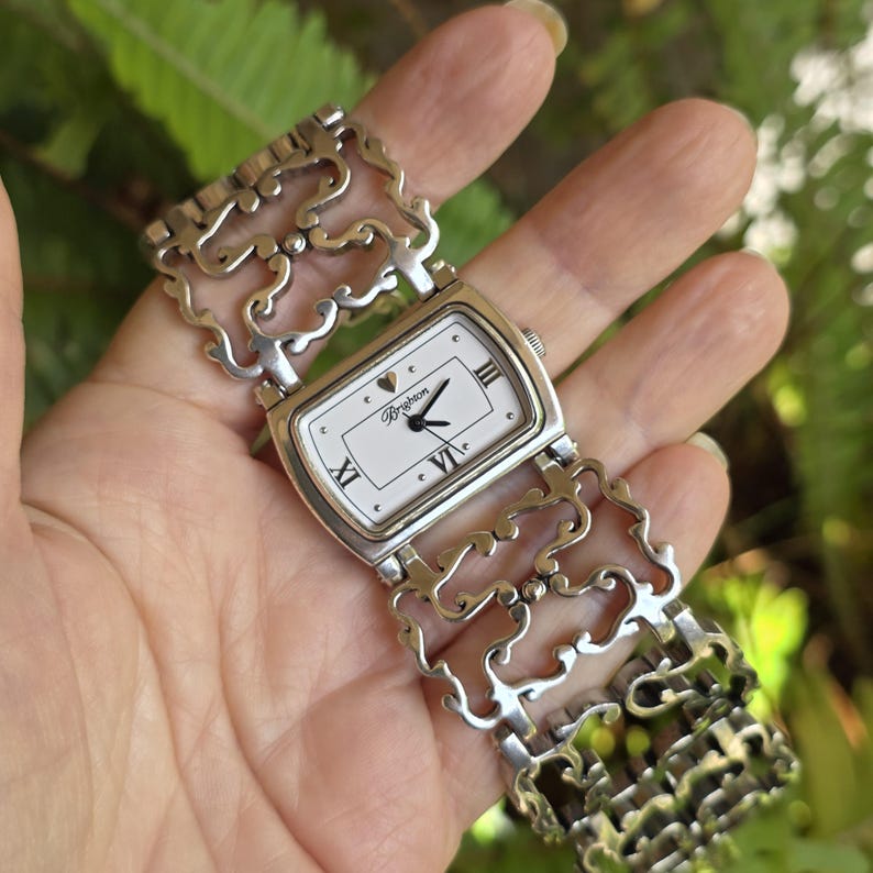 May include: Silver-toned Brighton watch with a rectangular white face. The watch features Roman numerals, a heart detail, and a decorative, open-link band. The watch is held in a hand against a blurred green background.