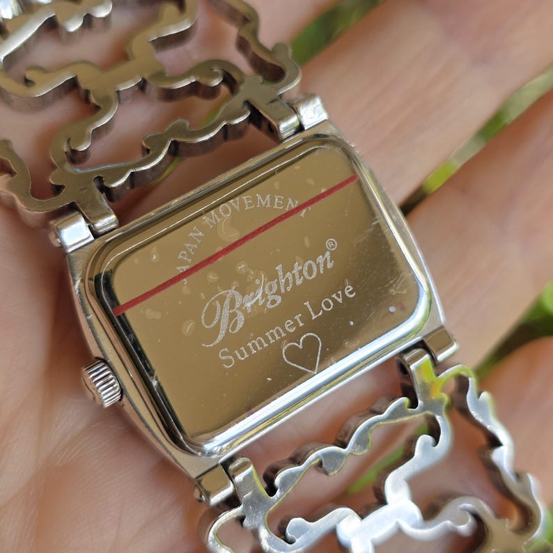 May include: A silver-toned Brighton watch with a rectangular face and an ornate, openwork band. The watch face is inscribed with "Brighton Summer Love" and a heart symbol. The words "Japan Movement" are also visible.