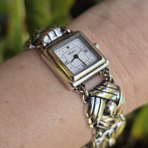 May include: A silver-tone analog wristwatch featuring a square white face with black hands and Roman numeral markers at the three, six, and nine o'clock positions. The watch band consists of interconnected, polished silver-tone metal links.