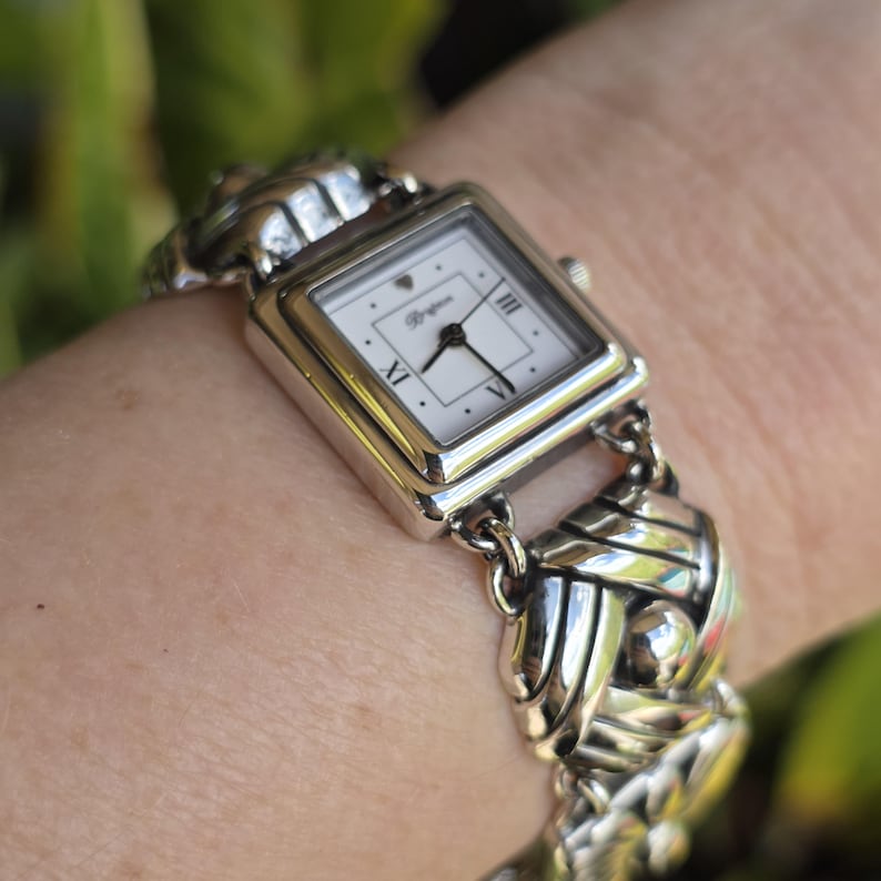 May include: A silver-tone analog wristwatch featuring a square white face with black hands and Roman numeral markers at the three, six, and nine o'clock positions. The watch band consists of interconnected, polished silver-tone metal links.