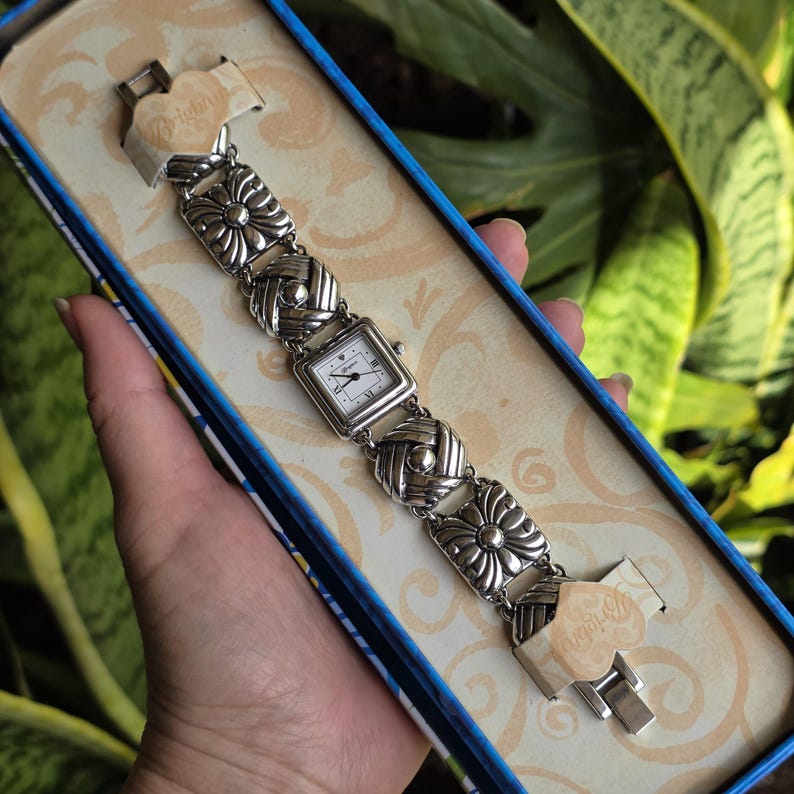 May include: A silver-tone analog watch with a square white face and black hands, featuring a link bracelet composed of alternating square floral-patterned and circular textured metal segments, presented in a branded box with heart-shaped tags.