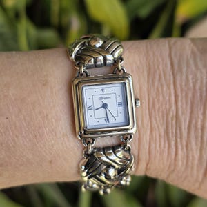 May include: A square-faced wristwatch with a white dial, black hands, and Roman numerals at the three, six, and nine o'clock positions. The watch features a silver-tone metal link bracelet with a braided, textured design and the Brighton logo.
