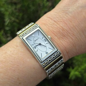 Op de afbeelding: Zilveren rechthoekig horloge met een witte wijzerplaat, zwarte cijfers en een klein hartje boven het Brighton-logo. De horlogeband is zilver- en goudkleurig, met een rekbaar ontwerp. Het horloge zit om een pols.