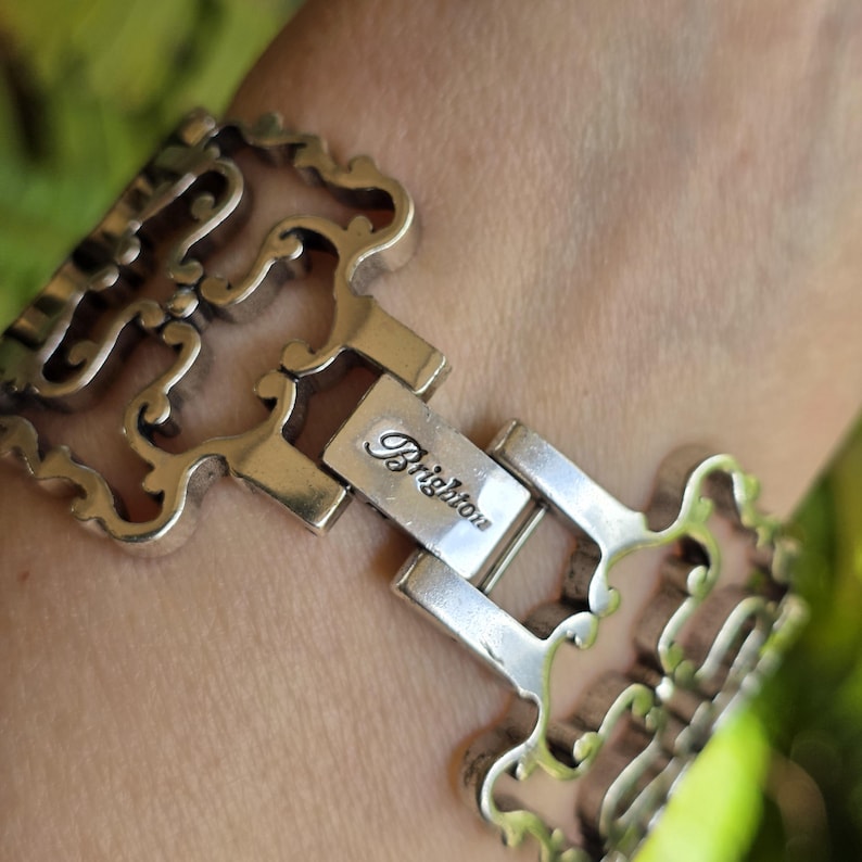May include: Silver-toned bracelet with an ornate, openwork design. The bracelet features a rectangular clasp with the word "Brighton" engraved on it. The intricate details and scrolling patterns give the bracelet a vintage aesthetic.