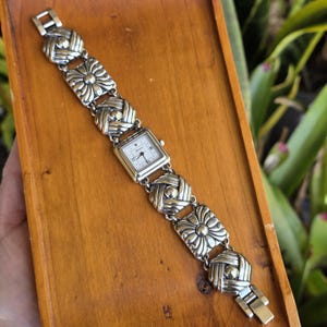 May include: A silver-tone wristwatch featuring a square white dial with black hands and markers. The band is composed of alternating square links with a floral-like embossed pattern and smaller, rounded woven-style links in a metallic finish.