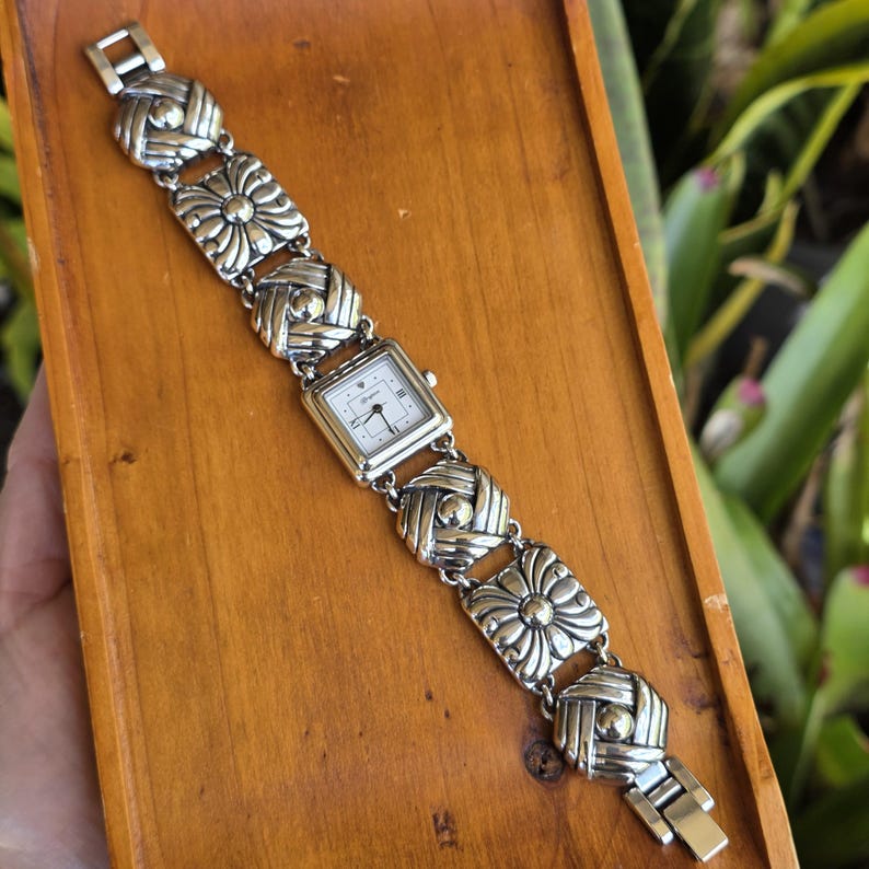 May include: A silver-tone wristwatch featuring a square white dial with black hands and markers. The band is composed of alternating square links with a floral-like embossed pattern and smaller, rounded woven-style links in a metallic finish.