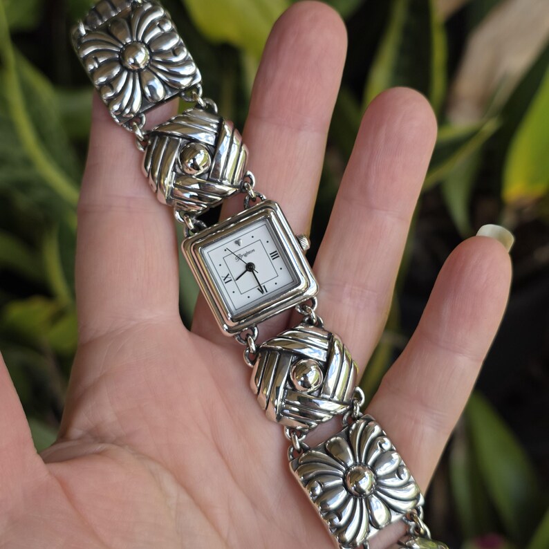 May include: A silver-tone link watch featuring a square white face with black hands and Roman numeral markers at the three, six, and nine o'clock positions. The band consists of alternating square links with embossed floral and woven patterns.