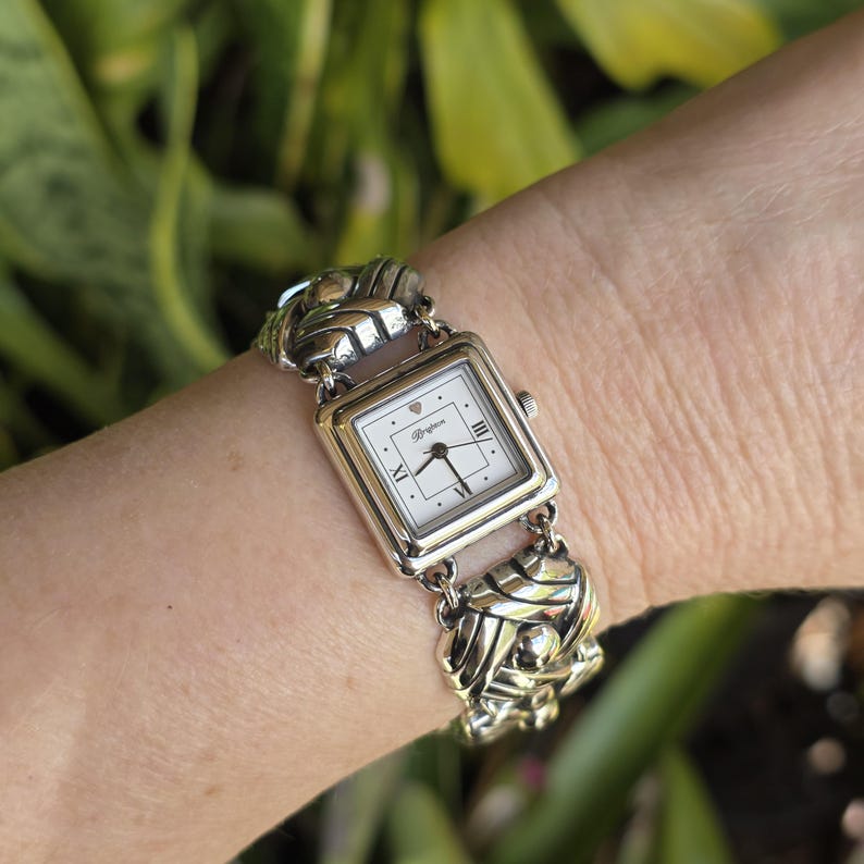 May include: A silver-tone wristwatch featuring a square white dial with black hands, Roman numerals at the three, six, and nine o'clock positions, and a small heart icon. The watch has a decorative, textured silver-tone metal link bracelet.