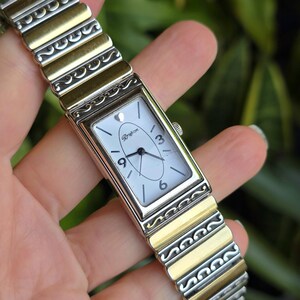 Op de afbeelding: Rechthoekig zilverkleurig horloge met een witte wijzerplaat, met de cijfers 3, 6 en 9. De horlogeband is een combinatie van zilver- en goudkleurig metaal. De wijzerplaat heeft de merknaam "Brighton". De lengte is ongeveer 17 cm.