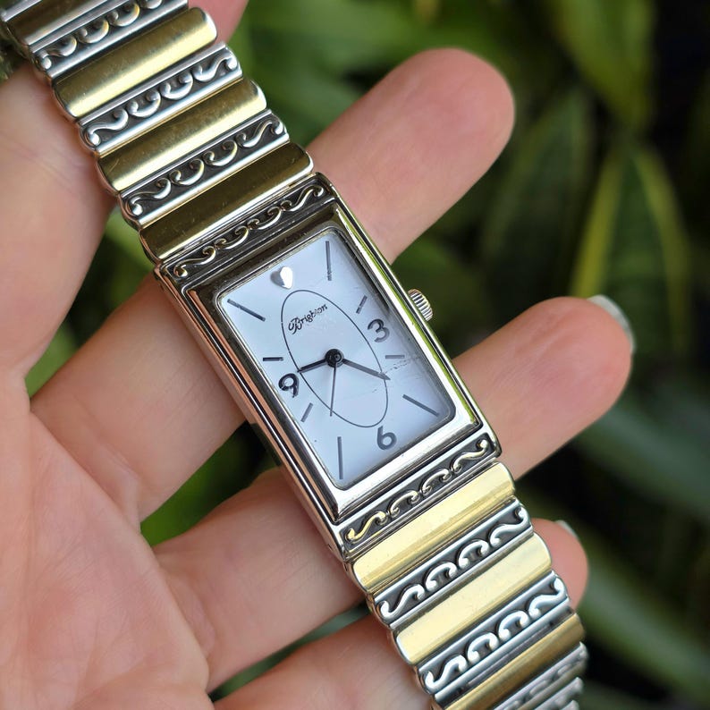 Op de afbeelding: Rechthoekig zilverkleurig horloge met een witte wijzerplaat, met de cijfers 3, 6 en 9. De horlogeband is een combinatie van zilver- en goudkleurig metaal. De wijzerplaat heeft de merknaam "Brighton". De lengte is ongeveer 17 cm.