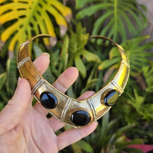 May include: A gold-toned collar necklace with three large, oval, black stones. The necklace has a geometric design with silver-colored accents. The background features green foliage.